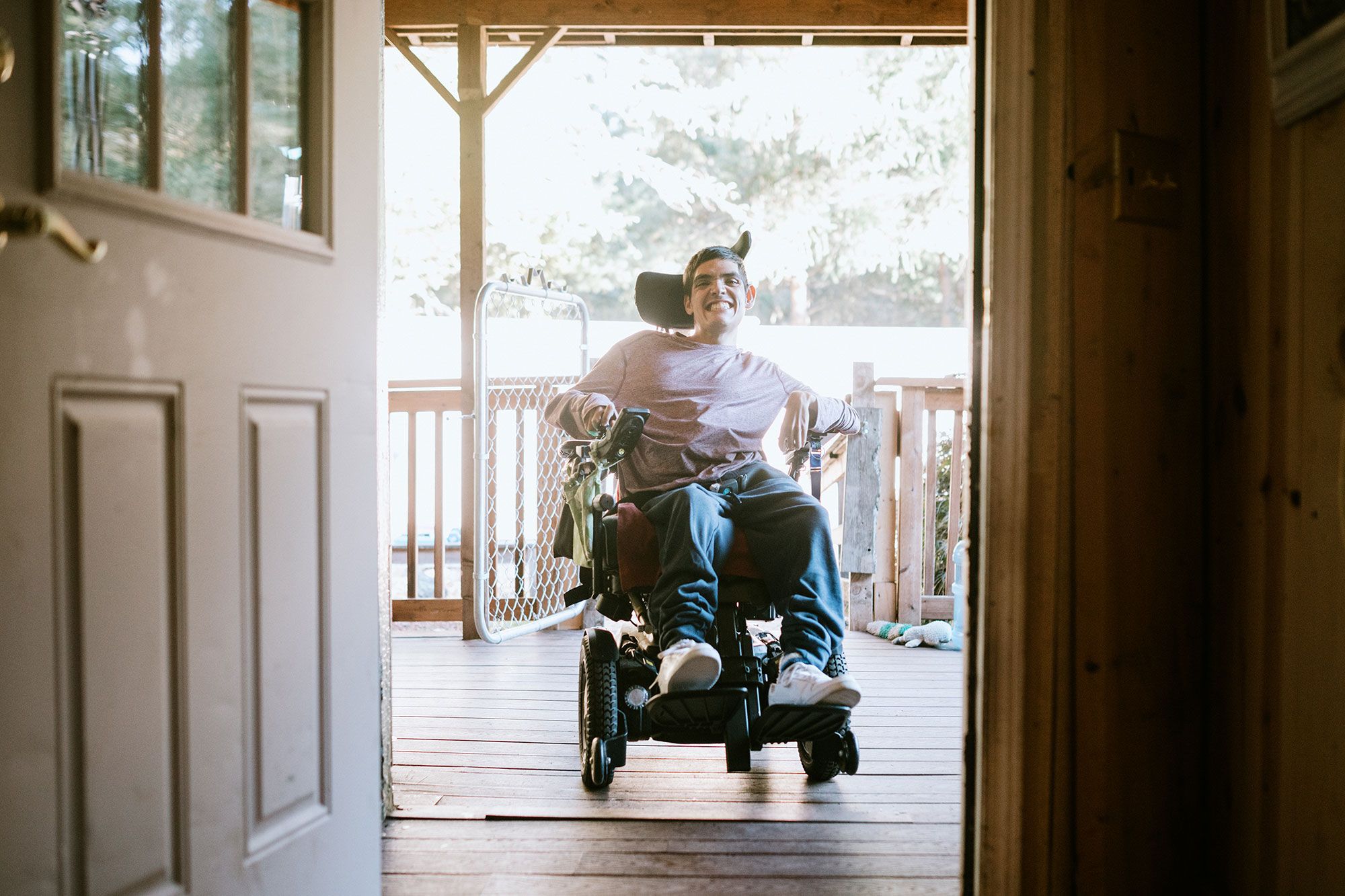 A teenage boy in a wheelchair on  a deck, smiling through an open door